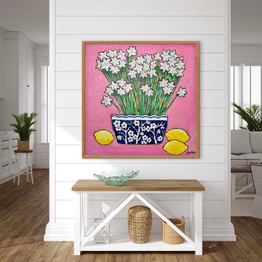 A whimsical painting of paperwhites in a blue and white floral vase surrounded by lemons against a bright pink background.