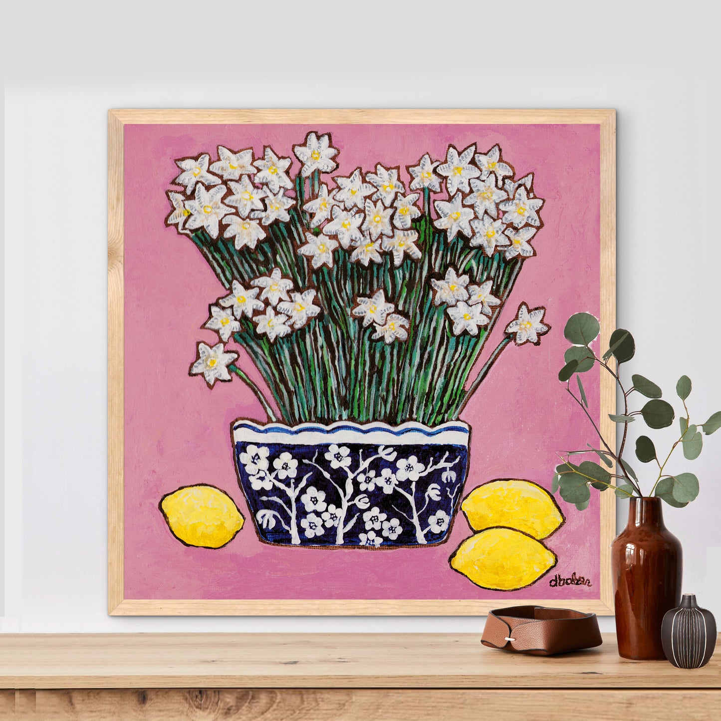 A whimsical painting of paperwhites in a blue and white floral vase surrounded by lemons against a bright pink background.
