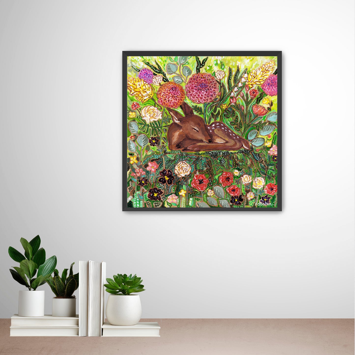 Framed artwork of a deer among flowers from Michigan Artist Deb Baker on a wall with decorative books and plants below. 