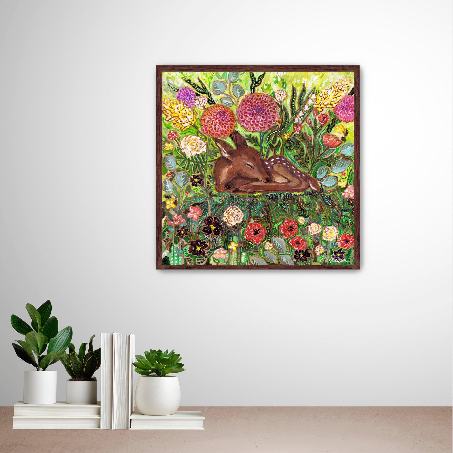 Framed artwork of a deer among flowers from Michigan Artist Deb Baker on a wall with decorative books and plants below. 